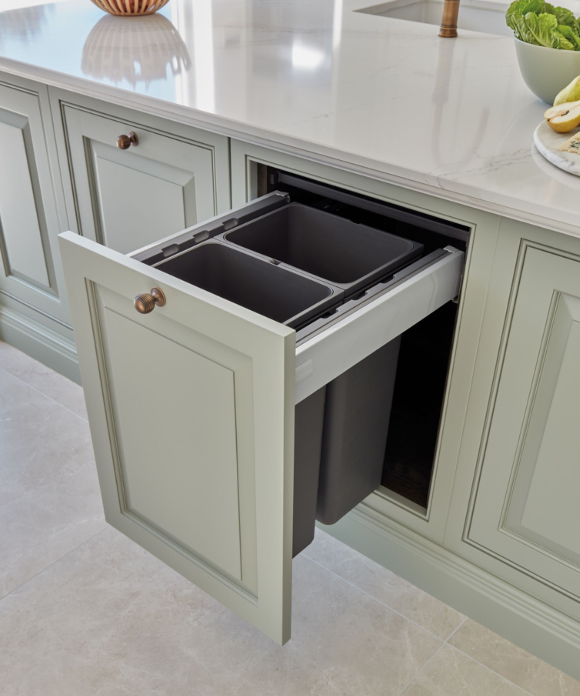 A built-in pull-out kitchen bin with two compartments, neatly hidden inside a pale green cabinet. Ideal for discreet waste and recycling storage beneath the countertop.