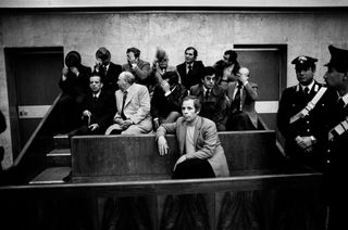 Black and white photograph of a man sat in a relaxed position in a court dock, flanked by authority figures. Several men are sat behind him attempting to hide their faces