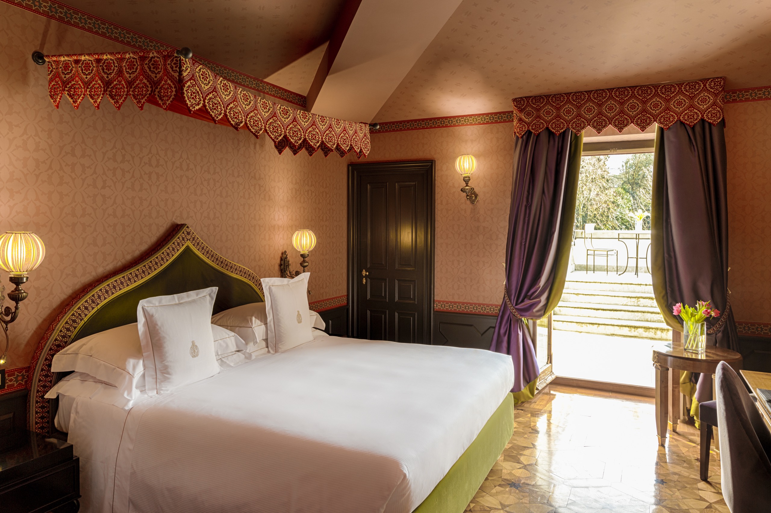 A sumptuously furnished hotel suite, featuring red and white, textured wallpaper, canopy-style bed curtains in red and gold, purple window curtains, an oriental-inspired velvet green bed, and softly glowing, rounded sconces.