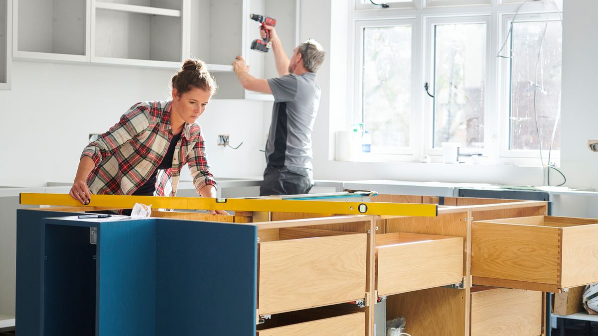 female using spirit level on bank of kitchen cupboard with male fitting cupboards on wall behind her