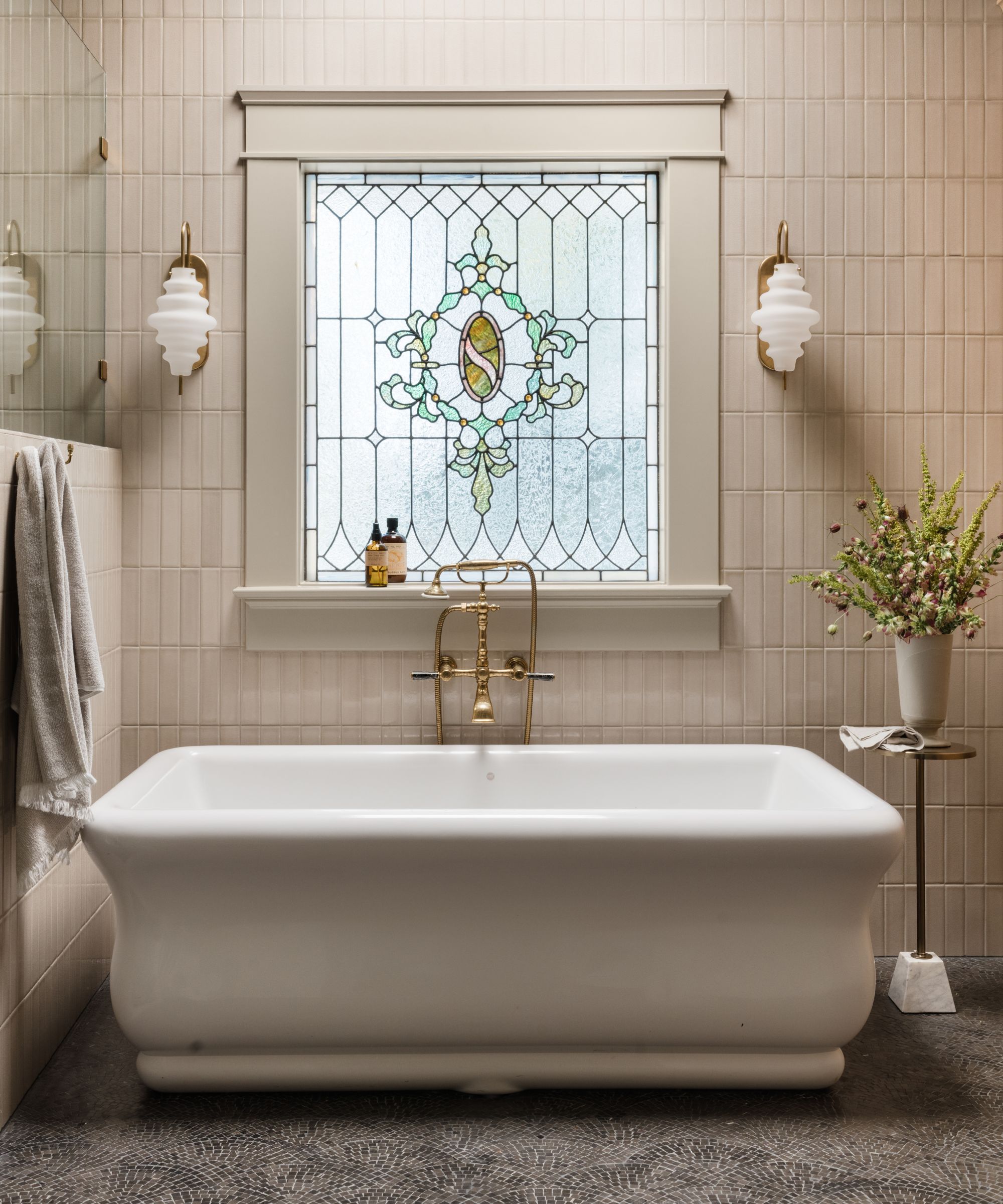 a large neutral primary bathroom with stain glass window, freestanding tub and sculptural sconces