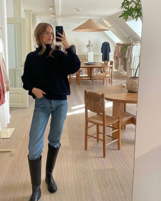 Clara wears a black turtleneck sweater, blue jeans and black knee-high boots.