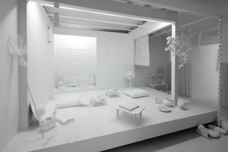 The Obliteration Room features various white pieces of art and furniture including a three small side tables with a tea pots on; a room fan; a fishing net; a cushion; a set of shelves with traditional Japanese plants.