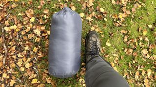 A sleeping bag in its pack, on the ground next to a foot to show the scale