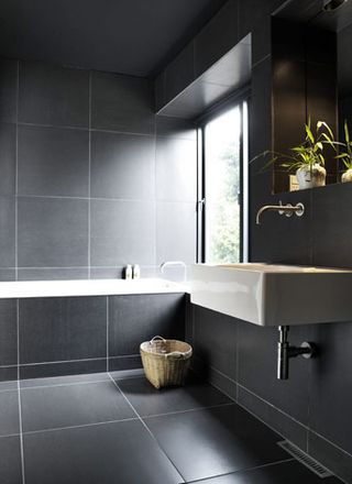 The bathroom is covered in large, dark grey tiles. To the right, is the sink area with a mirror above it. To the far wall is a bathtub with a window next to it.