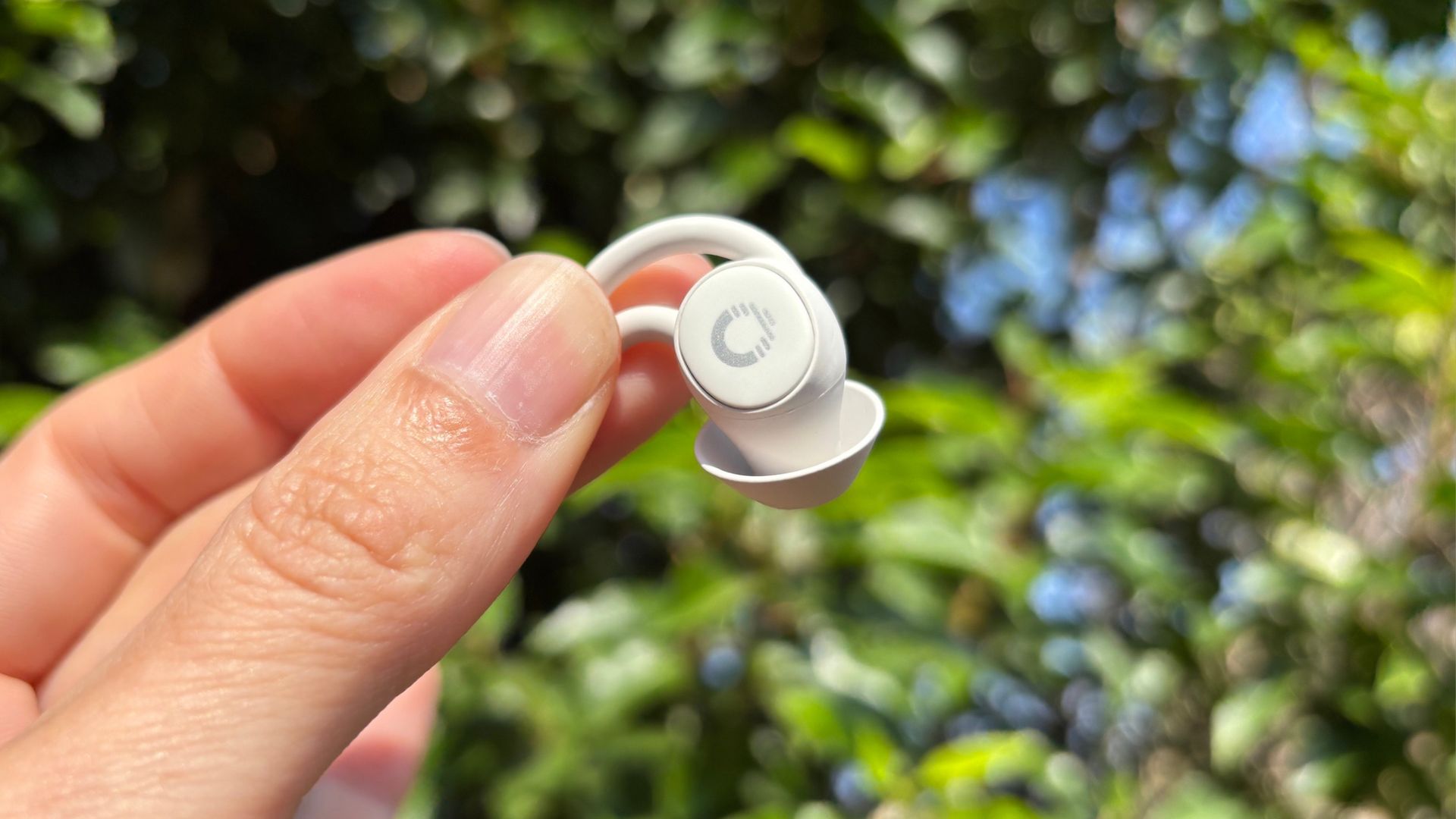 A hand holds a small white Ozlo Sleepbud 2 against a backdrop of a green plant