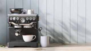 an espresso machine in front of pale blue wooden walls with a coffee cup inside it and milk jug at the side