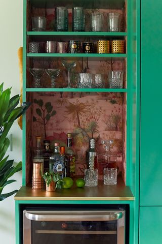 home bar in kitchen with green cupboards and shelves lined with tropical wallpaper and glasses on display
