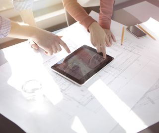 Plans of a house with and iPad on top and two people's hands pointing at different aspects of the design