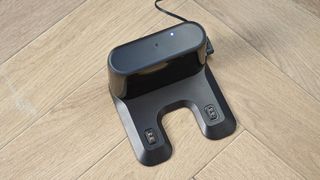 the charging station of a robot vacuum and mop in black by ultenic is photographed on a hardwood floor with a blue wall and a burnt orange terracotta and white striped rug