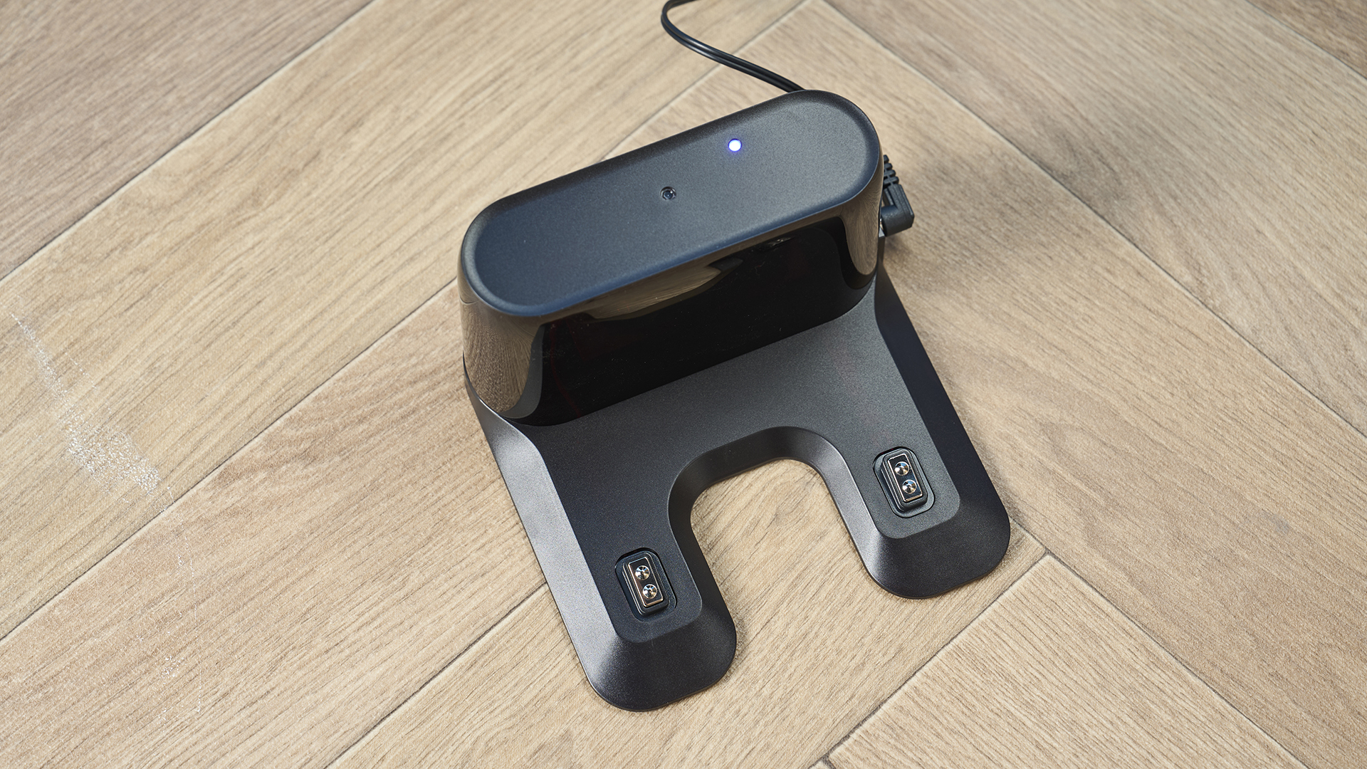 the charging station of a robot vacuum and mop in black by ultenic is photographed on a hardwood floor with a blue wall and a burnt orange terracotta and white striped rug