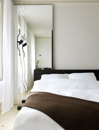 Focussing on a double bed with white linen and a brown throw thrown over the bed. To the left: A standing mirror and a standing lamp.