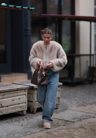 A woman wears a tan fur coat, Louis Vuitton bag, blue puddle jeans, and chestnut Ugg boots.