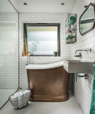A bathroom with a glass shower, a copper bath tub with a black window above it and a metal rack to the right of it, and a concrete sink with exposed pipes