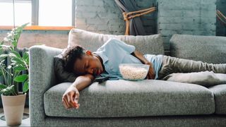 A man laying on grey sofa sleeping next to bowl of popcorn 