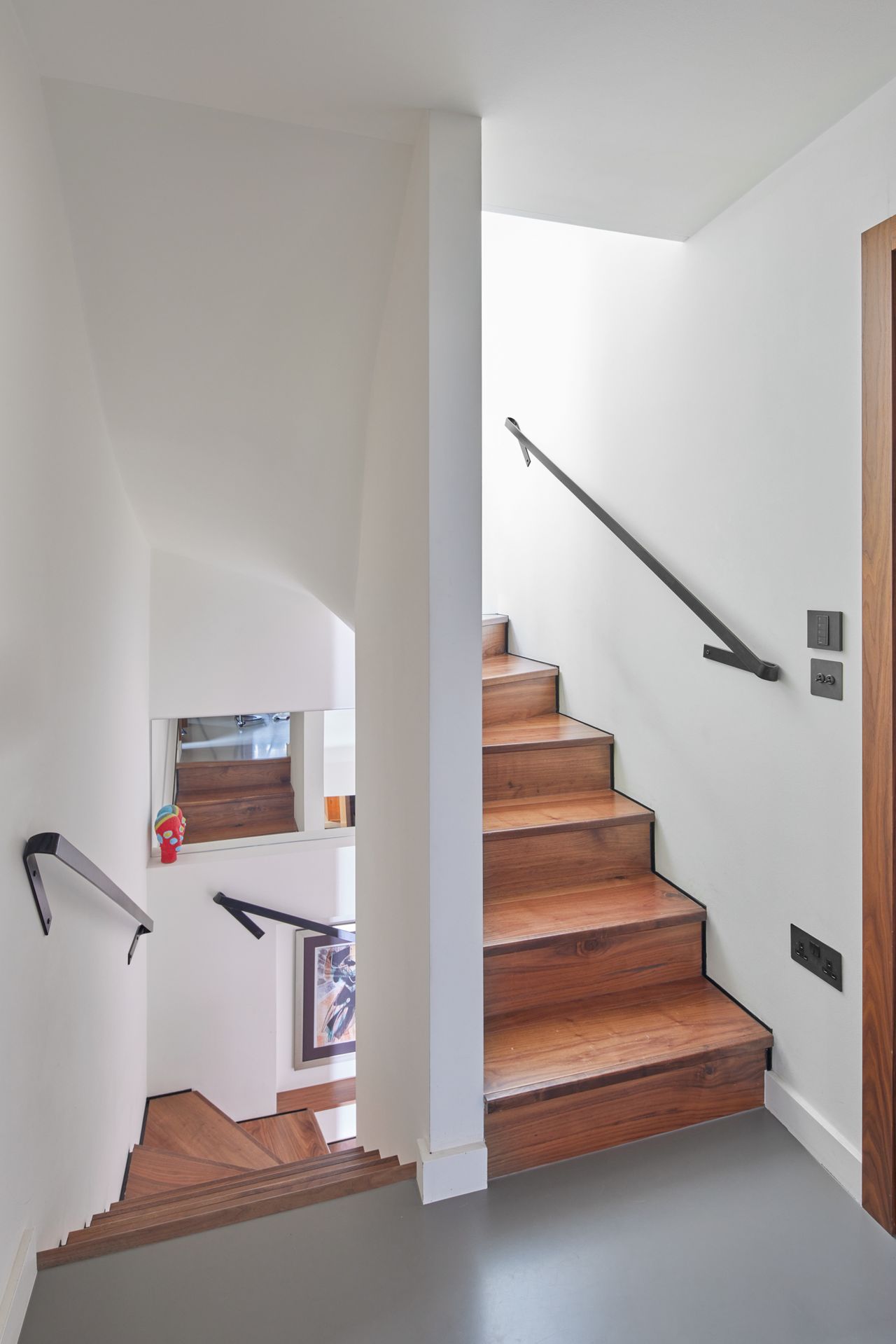 A modern wooden staircase ascending and descending from a small landing with a grey floor and white walls