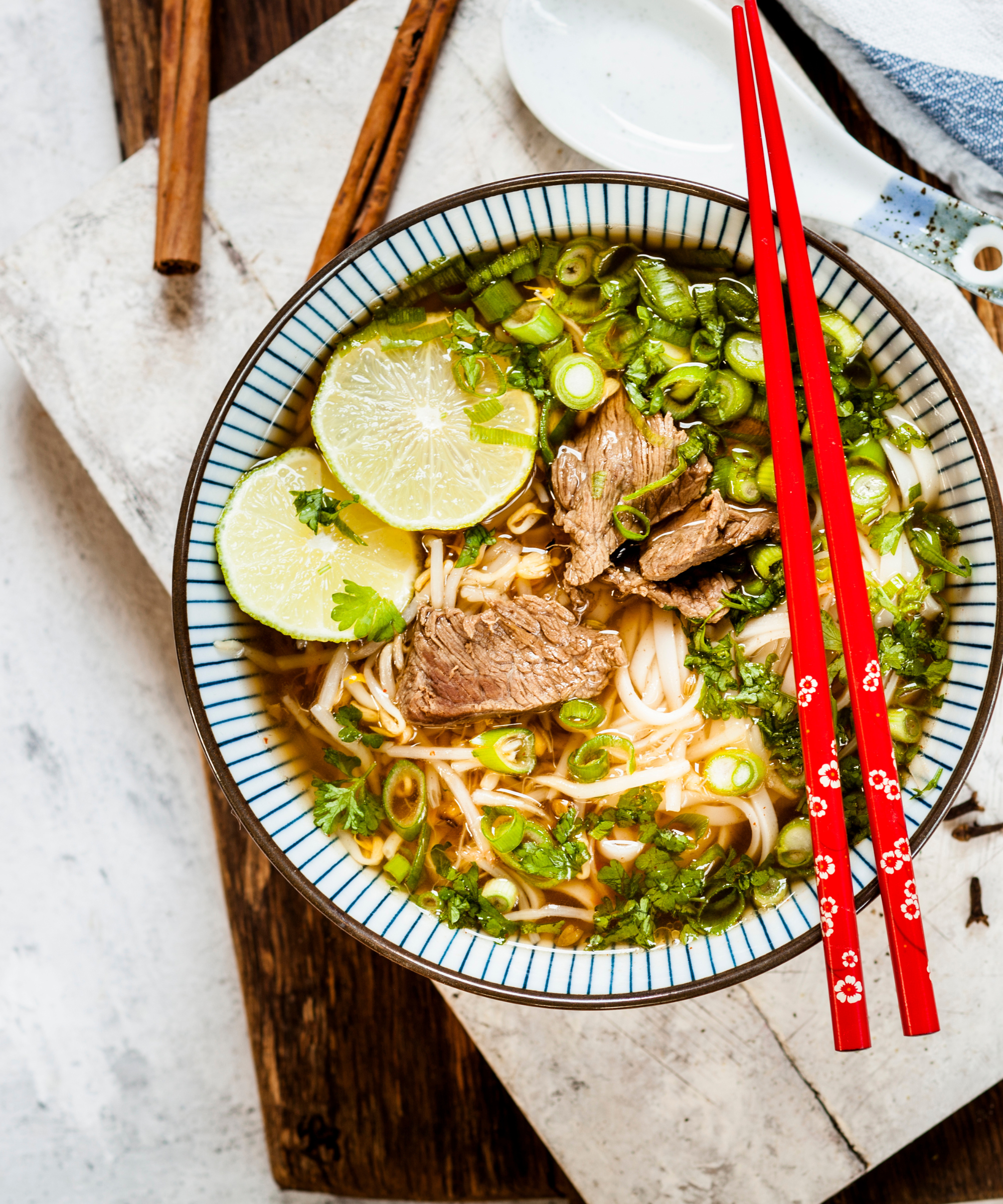 A bowl of beef phô noodles garnished with fresh lime and scallions, and red chopsticks resting on the bowl