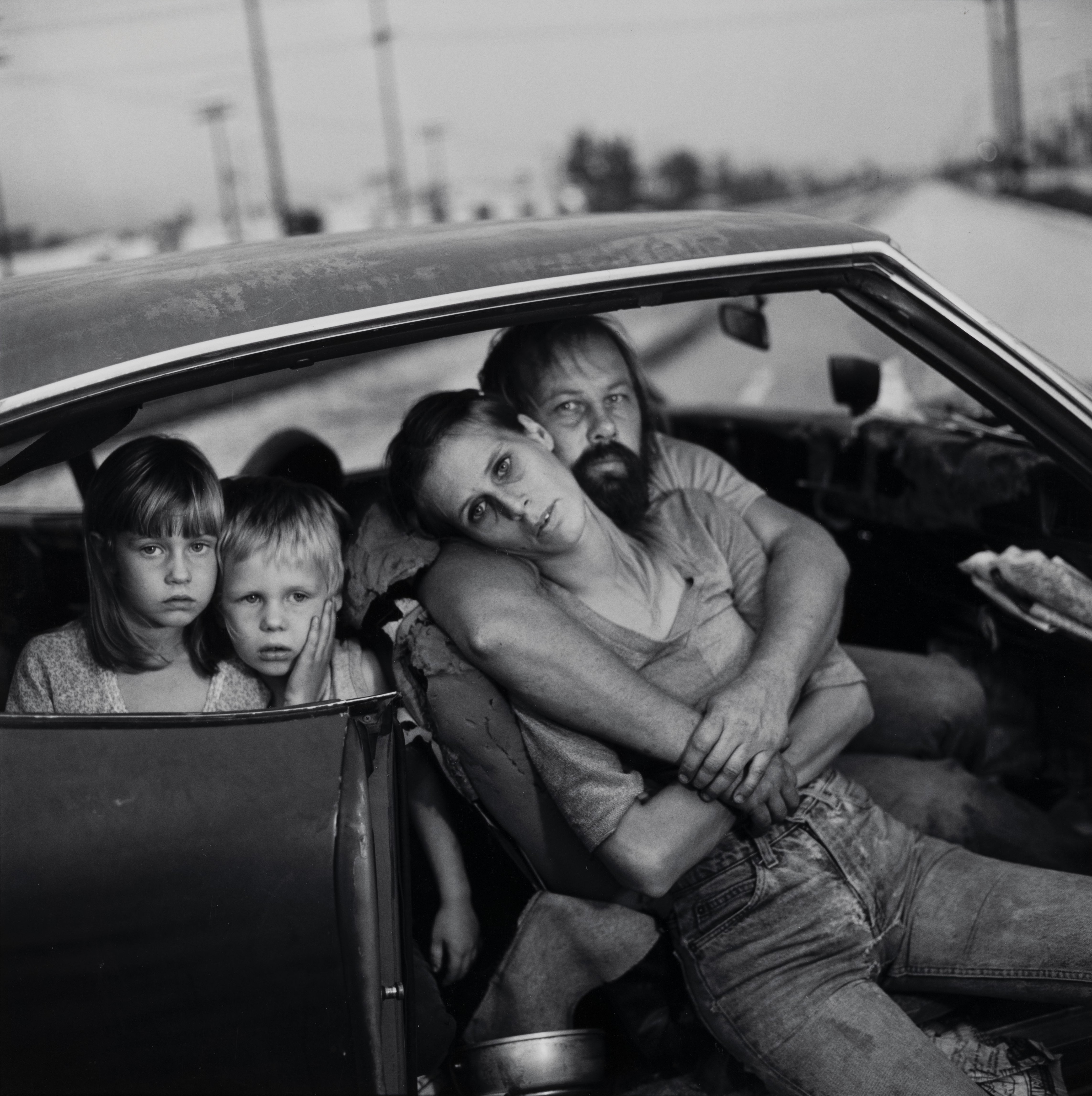 A woman leans exhausted against a bearded man in the front seat of a worn car, while two young children stare directly and blankly into the camera from the back.