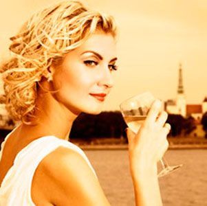 blond woman leaning on a fence in a field drinking a glass of wine