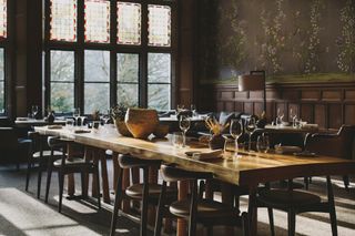 A modern rustic restaurant in the countryside is decorated with mid-century wood and leather furniture with a central, huge dining table, floral wallpaper, and stained-glass windows.