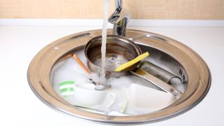 A stainless steel pan soaking in soapy water in a sink with teacups, drinking glasses and cutlery