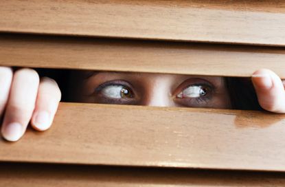 A tearful young woman with bloodshot eyes peers suspiciously to the side through the slats of a wooden venetian blind.