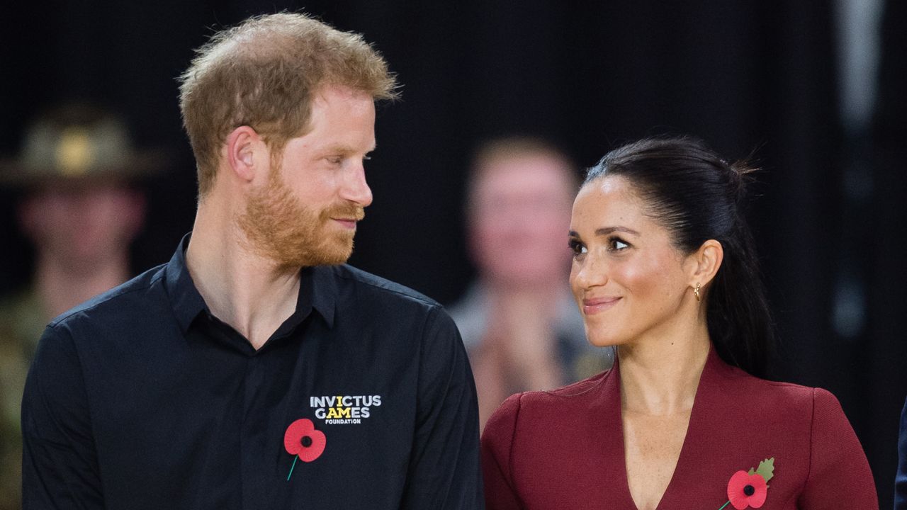 Prince Harry wears a black Invictus Games polo shirt and gazes adoringly at his wife, Meghan Markle, who is wearing a dark red V-neck top