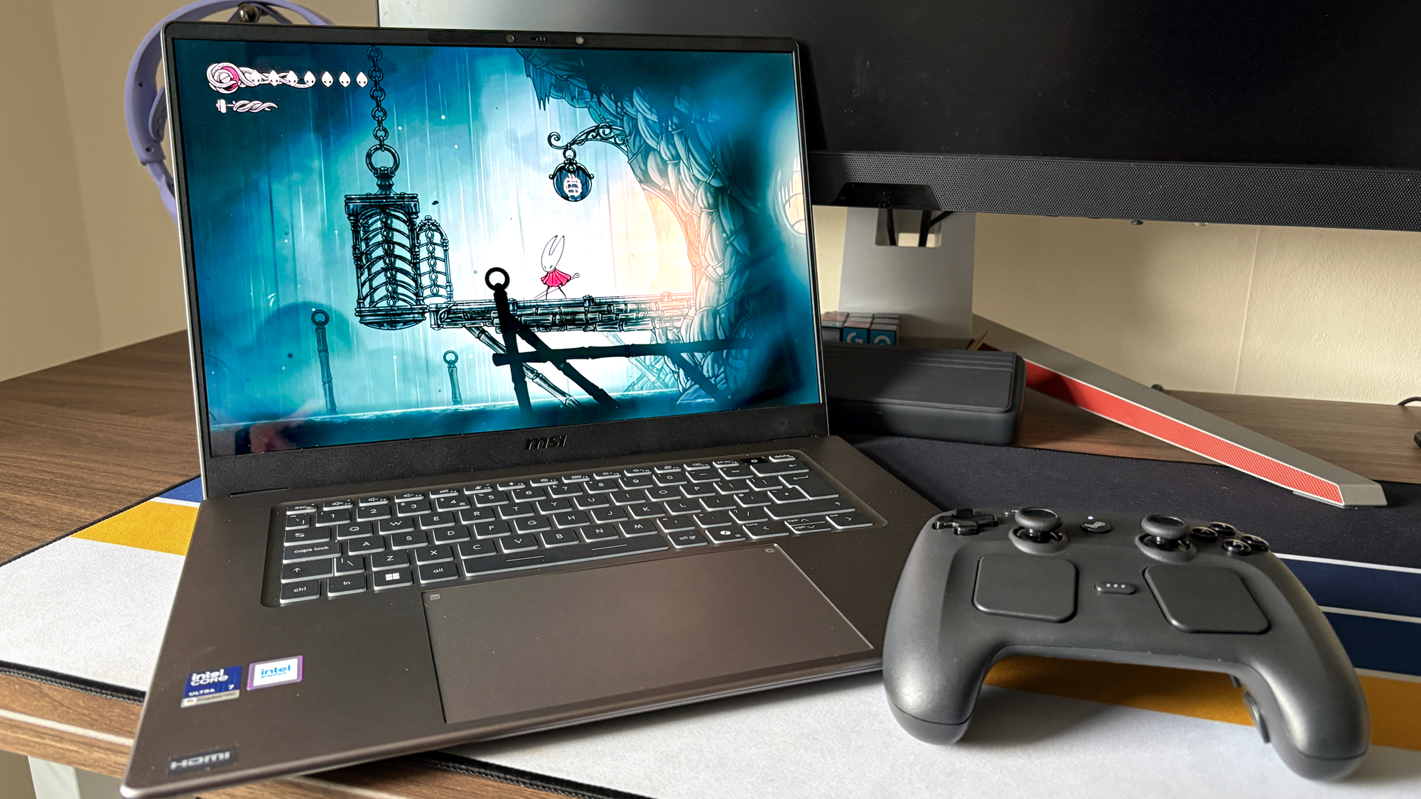 MSI Prestige 16 AI+ on table showing Hollow Knight Silksong on screen with controller next to it