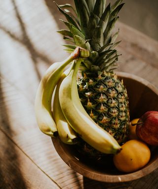 A wooden fruit bowl with a pineapple, bananas, apples, and oranges on a wooden surface