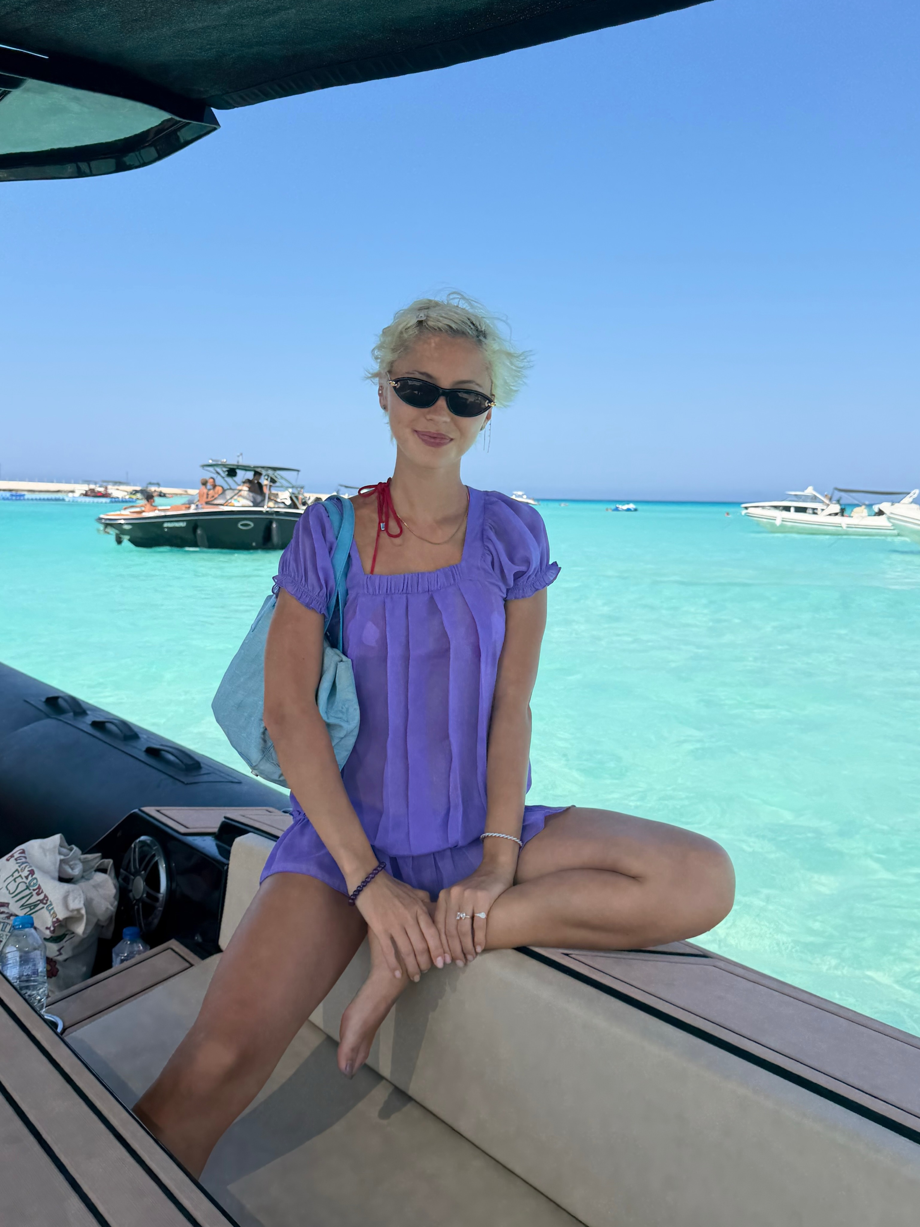 Iris Law sits on a boat wearing a purple minidress with a turquoise bag.