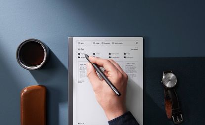 A hand holding a stylus above a tablet next to a coffee cup, a watch and a wooden eye glasses case.