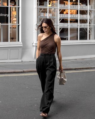 British influencer Jessica Skye walks across a London street wearing aviator sunglasses, a brown one-shoulder top, black trousers, a cream Loewe puzzle bag, and brown strappy flat sandals