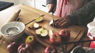 a person using a wooden chopping board to chop apples, with baking ingredients surrounding the kitchen countertop