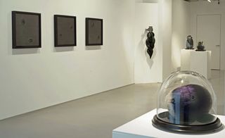 Three dark pictures hang on one wall, with a black, interwoven light fixture and a dark object under a glass dome in the foreground