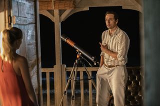 Neville (Ralf Little) stands on the veranda of his beachfront shack at night, next to a telescope. In the foreground, Sophie (Chelsea Edge) stands with her back to the camera, out of focus