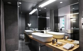 Bathroom, grey stone wall tiles, white bath, wooden unit with white sink on top, wall mirror behind sink, strobe light above sink, spotlight in ceiling , white towels