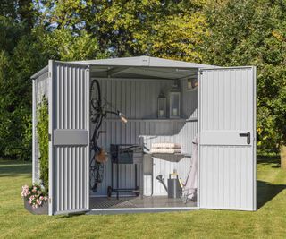 A light grey metal garden storage shed from Hörmann set in a garden. The doors are open and a bike, bbq and garden furniture are stored inside.