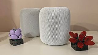 HomePod 2 in white on a white table, with original HomePod in white behind it. Two Lego flowers sit at the base