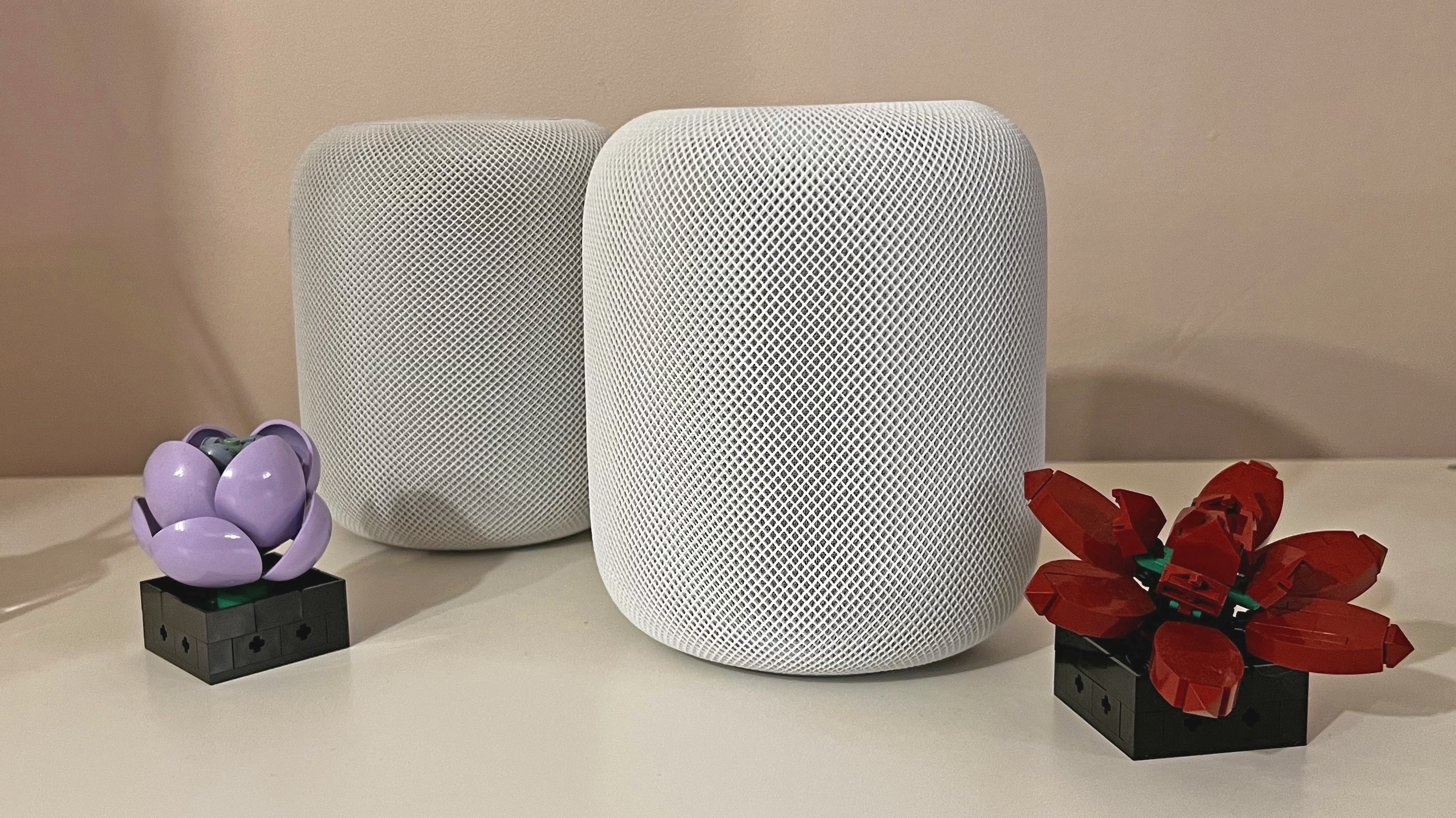 Apple HomePod 2 in white on a white table, with original HomePod in white behind it. Two Lego flowers sit at the base