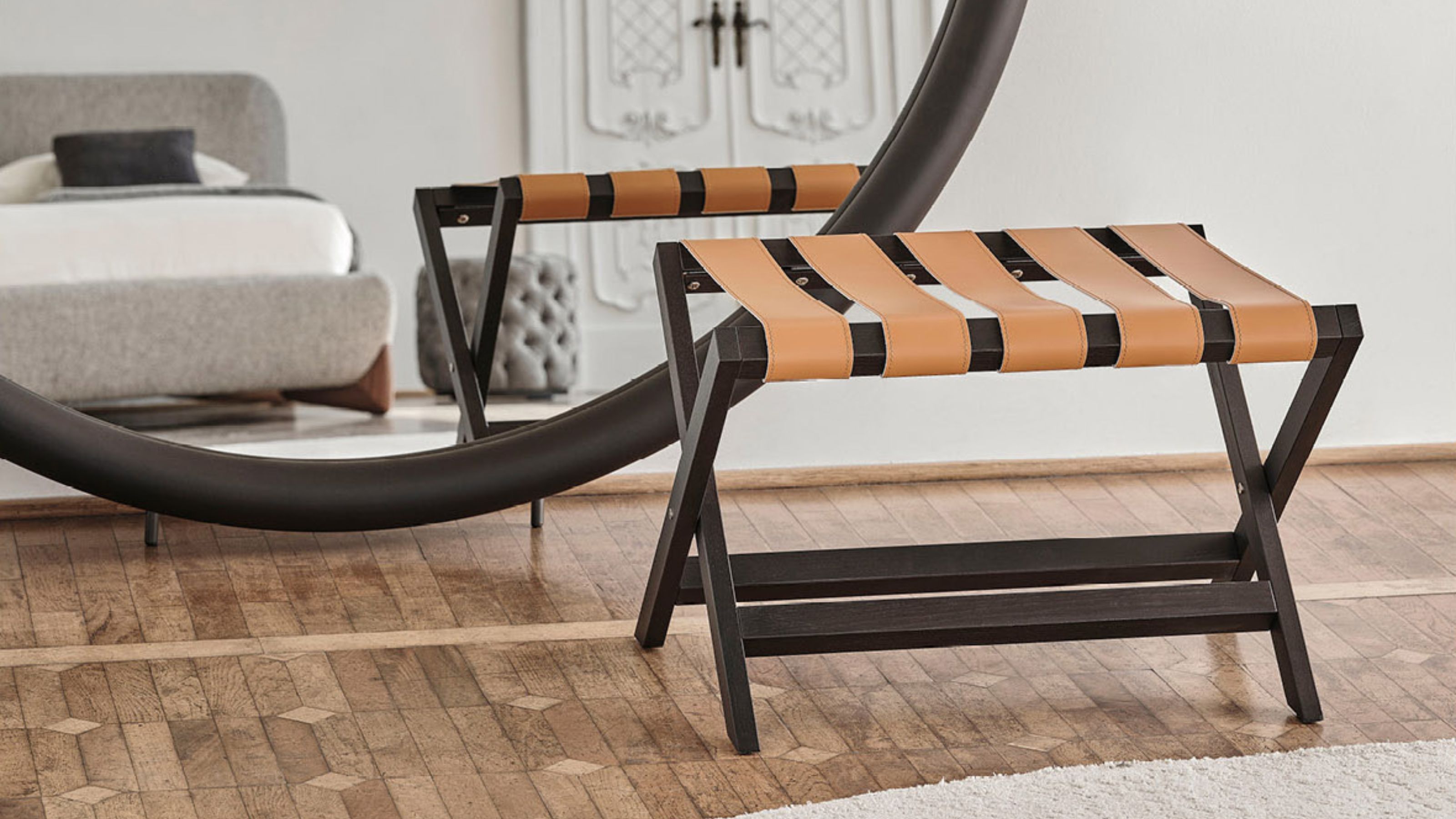 Porada portavaligie luggage rack in bedroom with large round black mirror, timber floor, gray bedframe, white sheets, white wall, and ornate white doors