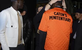 Man fixing a hat on a model wearing an orange vest with Caten's Penitentiary printed on the back