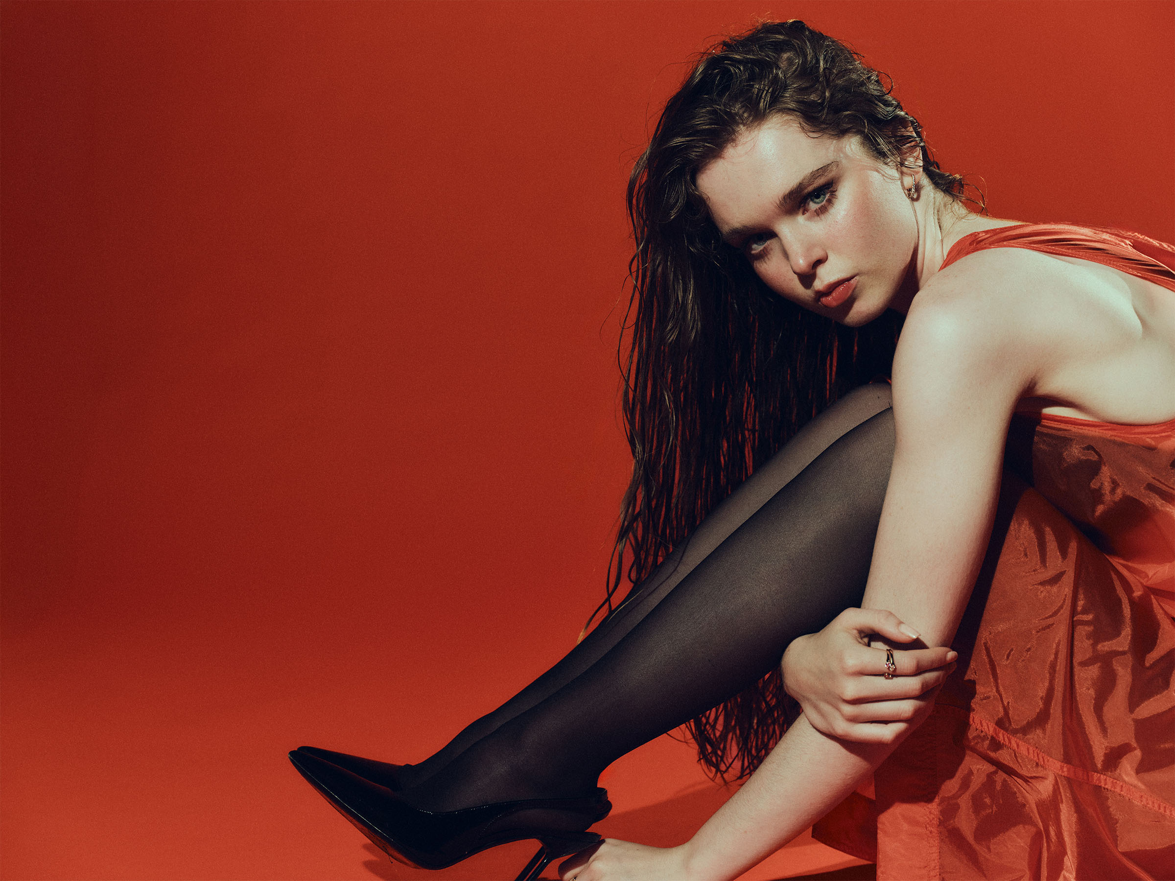 Carrie actress Summer Howell photographed by Jonny Marlow. Her hair is wet, and she is sitting down leaning over her legs, wearing a red dress, black tights, and black pumps.