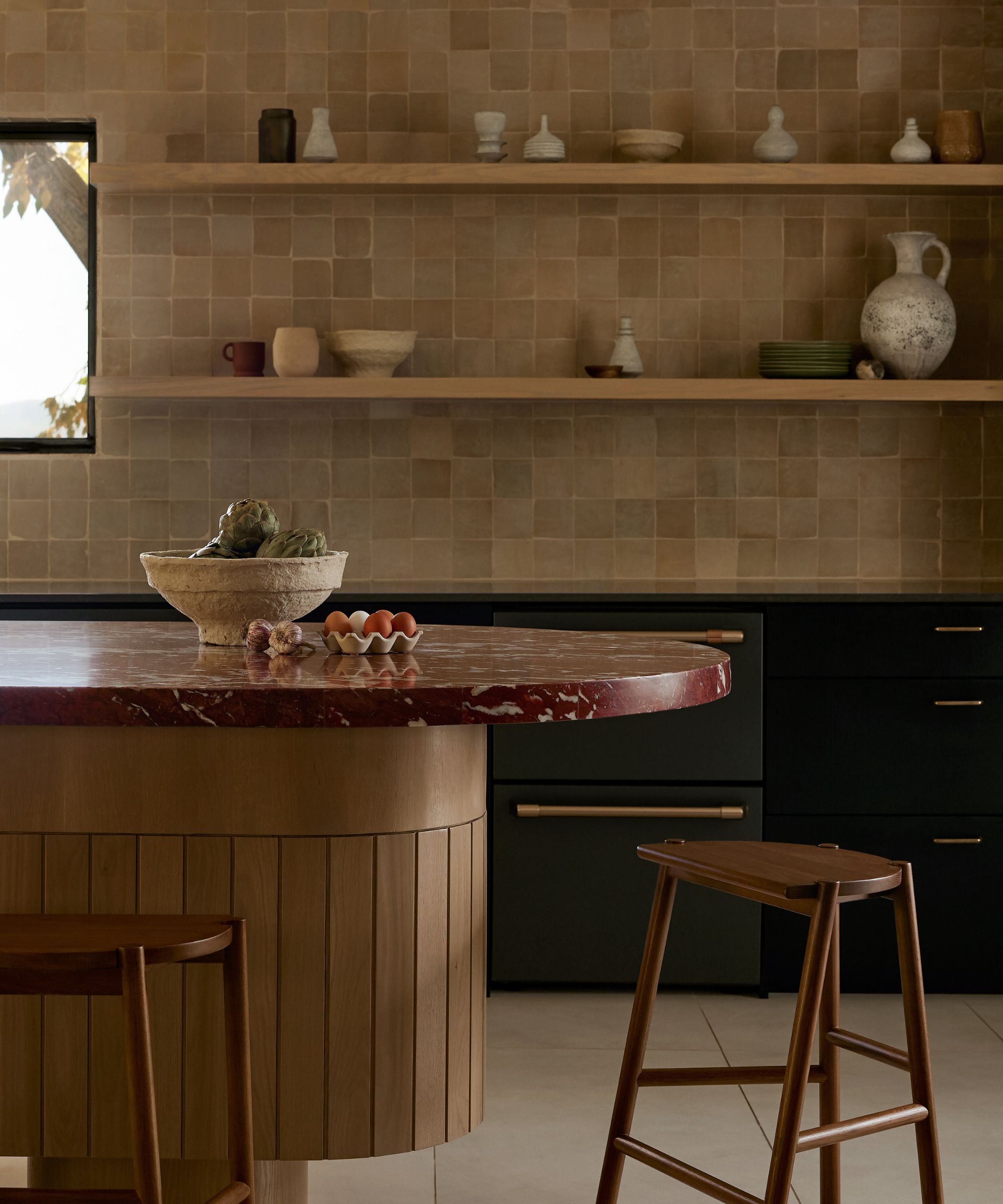 A kitchen with beige square tiles on the wall with two wooden open shelves on top, black countertops, and a curved wooden island topped with a burgundy marble countertop
