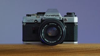 Olympus OM-10 film camera, with a Zuiko 50mm f/1.8 lens, on a wooden surface in front of a lilac background