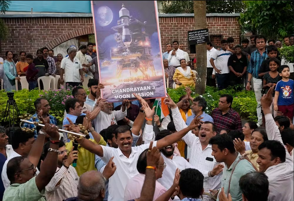 A group of cheering, smiling people hold signs depicting the Chandrayaan-3 lander.