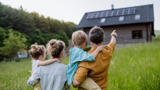 family comprising male, female adults and male and female children stood in field with backs to camera pointing at timber clad house with solar panels on roof