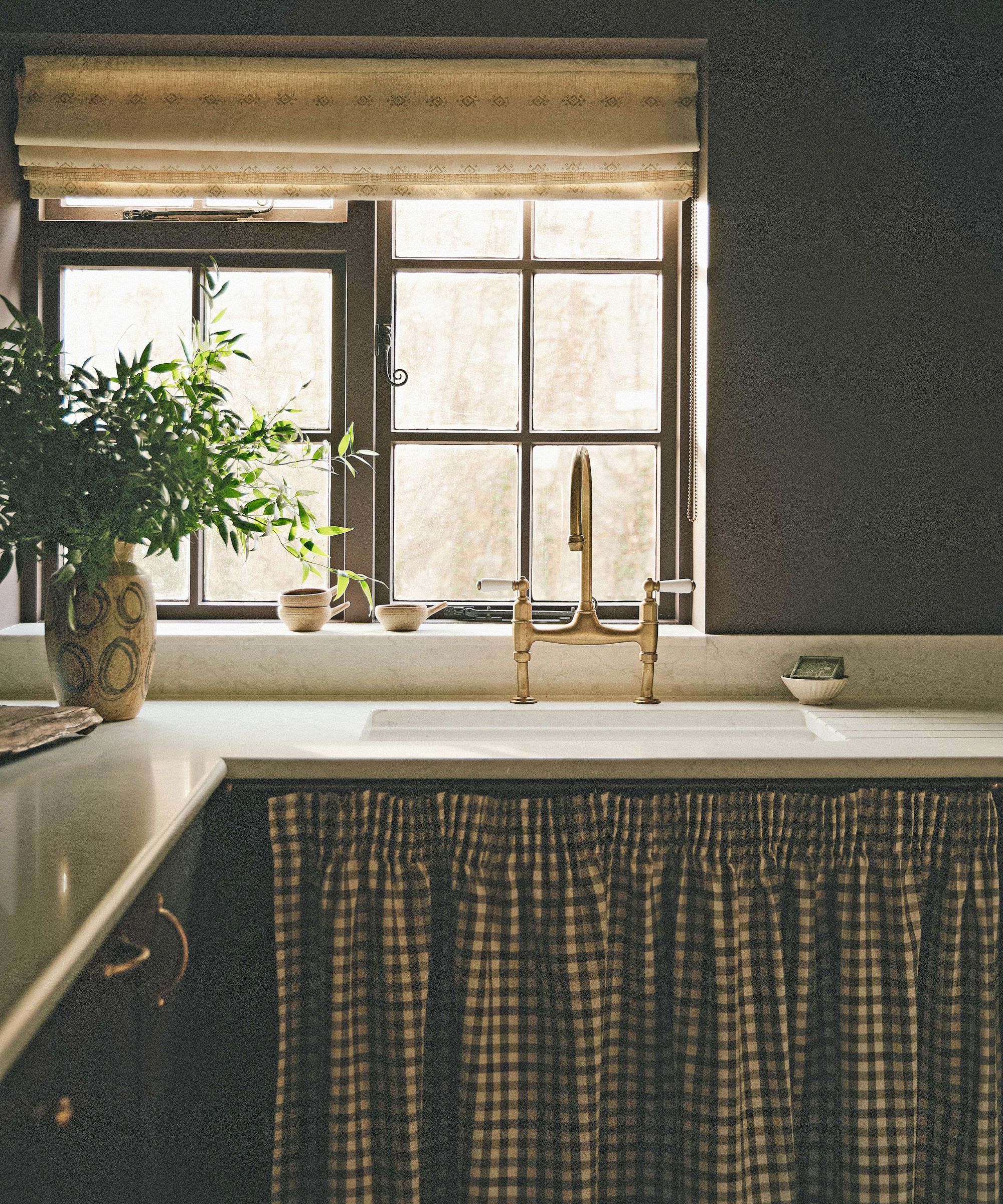 a dark olive green brown pantry in a cottage home with sink, window and gingham cabinet skirt