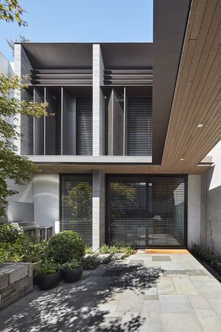 The exterior of a grey 2 floors residential home with full lenght windows featuring grey open blinds. Grey concrete tile floor with potted green plants