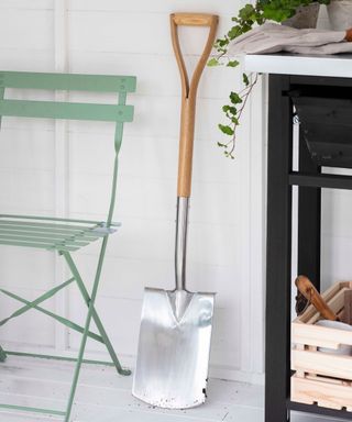 a wooden handled spade leaning against a white timber wall in a shed beside a pale green folding metal bistro chair