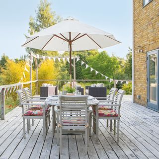 An alfresco dining area with outdoor dining set furniture, cream-coloured parasol on decked floor, and bunting decor hung around perimenter of deck